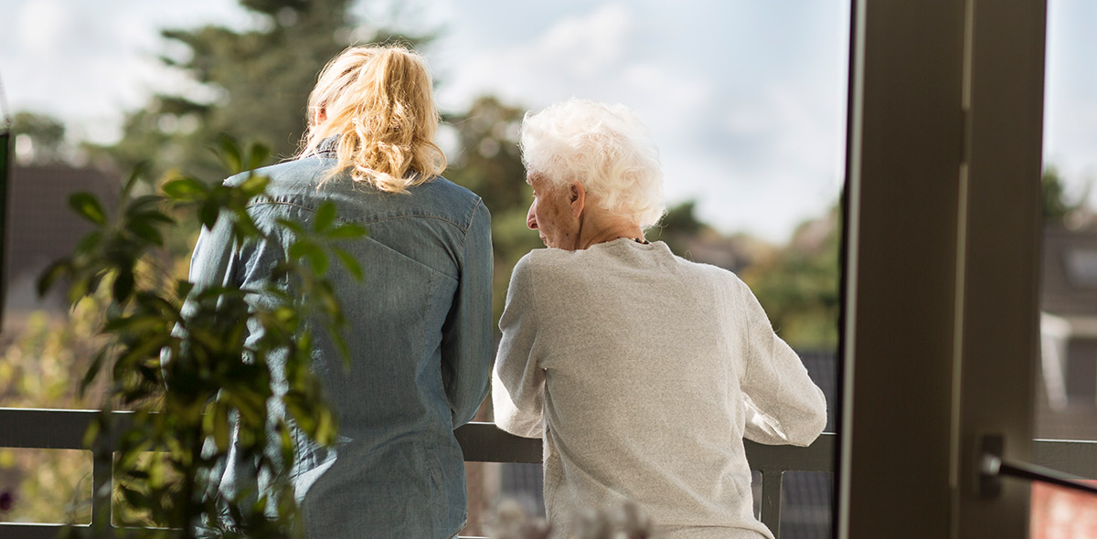 Afbeelding van een oudere vrouw die met haar dochter op het balkon staat naar buiten te kijken